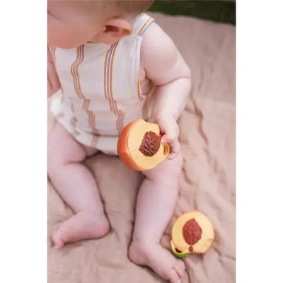 Chewable toy shaped like a peach held by a baby, with a second toy on the ground, set on a textured surface.