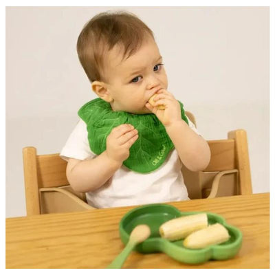 Child in a high chair wearing a green leaf-shaped bib, eating banana slices from a green kale-shaped plate and spoon set.