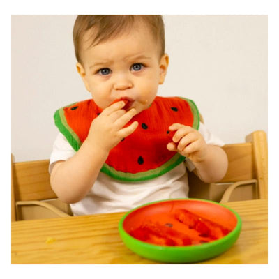 Infant wearing a watermelon-themed bib, eating watermelon from a green plate, sitting at a wooden table.