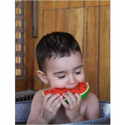 A young child in a bath is playfully chewing on a colorful chewable toy shaped like a watermelon, Wally The Watermelon.