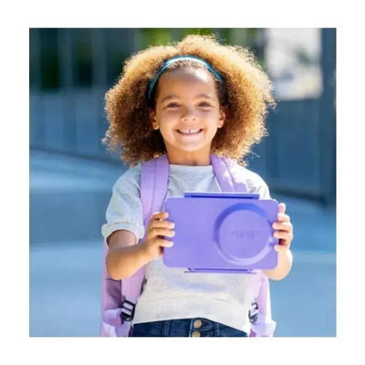 Smiling child holding a Galaxy Purple OmieBox Bento Box, wearing a gray shirt and backpack outdoors.