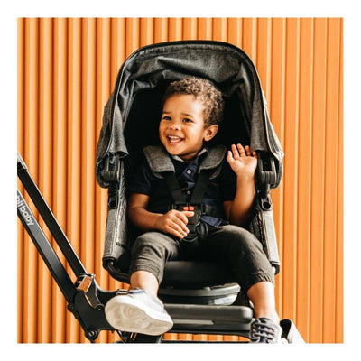 Child smiling and waving while seated in an Orbit Baby G5/X5 Helix+ seat with a gray canopy against an orange backdrop.