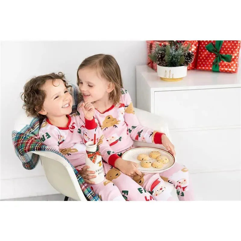 Two children in festive pajamas sit together, sharing a laugh with a plate of cookies and a milk bottle for Santa.