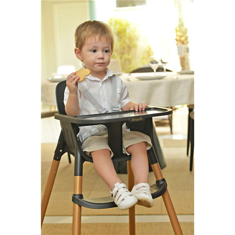 Child sitting in a Peg-Perego high chair, wearing a white shirt and holding a cookie in a dining setting.