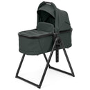 Sage green Peg Perego Ypsi bassinet with a black stand, designed for infants, featuring a removable canopy and padded interior.