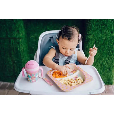 Baby seated in a high chair, using a bamboo fiber spoon and fork to eat food from a colorful plate with fruit.