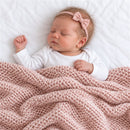 A light pink knit blanket drapes over a sleeping baby with a pink bow headband, resting on a white surface.