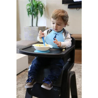 A toddler in a high chair wears a blue silicone baby bib while eating from a bowl with a spoon.