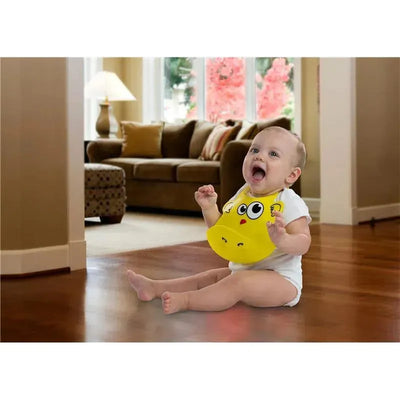 Baby sitting on hardwood floor wearing a yellow silicone bib with a cartoon face, smiling happily in a cozy living room.