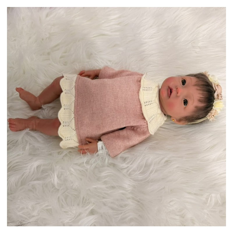 Reborn baby doll with African American features, wearing a pink dress and a floral headband, resting on soft white fabric.