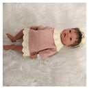 Reborn baby doll with African American features, wearing a pink dress and a floral headband, resting on soft white fabric.