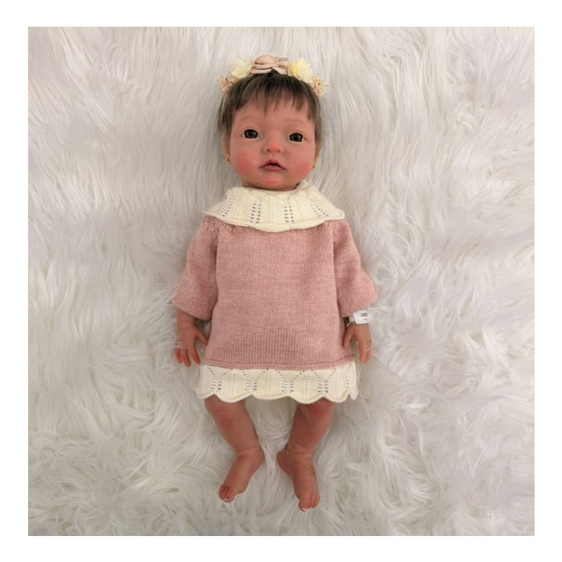 Reborn baby doll with African American features, wearing a pink knitted dress and floral headband, on a fluffy white surface.