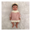 Reborn baby doll with African American features, wearing a pink knitted dress and floral headband, on a fluffy white surface.
