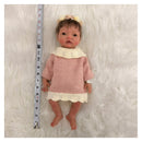 African American silicone reborn baby doll wearing a pink sweater dress with a white collar, measuring tape alongside.