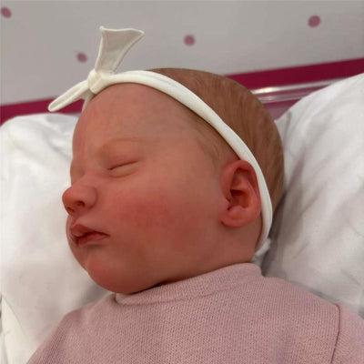 Asleep reborn baby doll with white vinyl skin, wearing a pink shirt and a white headband, resting on a soft surface.