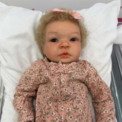 Reborn baby doll named Shyann with curly hair, wearing a pink floral outfit, resting on a white pillow.