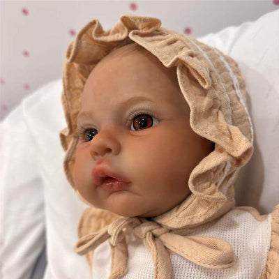 Reborn baby doll with realistic features, wearing a beige bonnet and white outfit, resting on a soft surface.