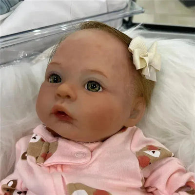 Reborn baby doll with light brown hair and green eyes, wearing a pink outfit with animal prints, adorned with a bow.