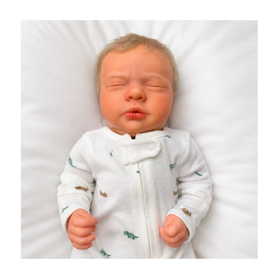 Reborn baby doll named Mark, crafted from white vinyl, wearing a printed onesie and resting on a soft surface.