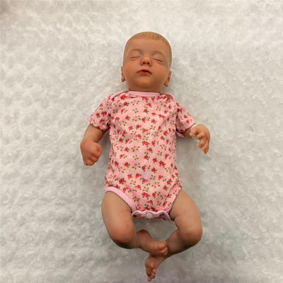 Reborn baby doll named Marnie 2, made of white vinyl, wearing a pink floral onesie, lying on a textured white background.