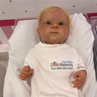 Reborn baby doll named Bettie, featuring white vinyl skin, rooted blonde hair, and blue eyes, dressed in a white shirt.