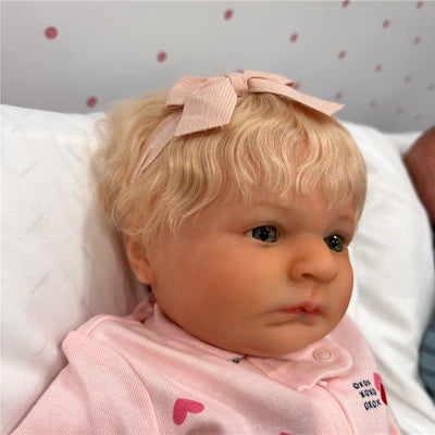 Reborn baby doll with light blonde hair and a pink bow, dressed in a pink top with hearts.
