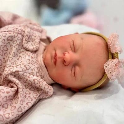 Reborn baby doll, Aria, with closed eyes, wearing a heart-patterned outfit and a pink lace bow headband.