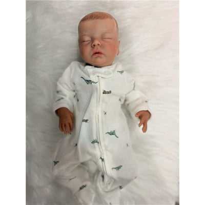 Reborn baby doll in a white dinosaur-printed onesie, resting on a soft white background.