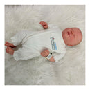 Reborn baby doll made of white vinyl, wearing a white outfit with a hospital wristband, lying on a fluffy surface.