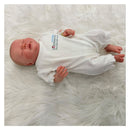 Reborn Baby Doll with a lifelike appearance, dressed in a white outfit featuring embroidered text, resting on a fluffy white surface.