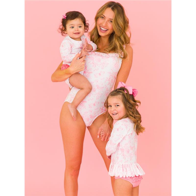 A woman holds a baby and stands beside a girl, all wearing matching pink butterfly-themed swimsuits against a pink background.
