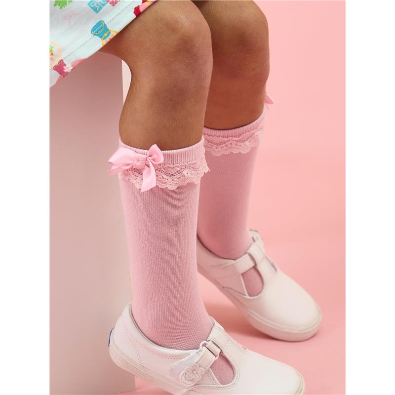 Knee-high pink socks with lace trim and bows, worn by a child with white shoes, against a pink background.