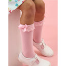 Knee-high pink socks with lace trim and bows, worn by a child with white shoes, against a pink background.