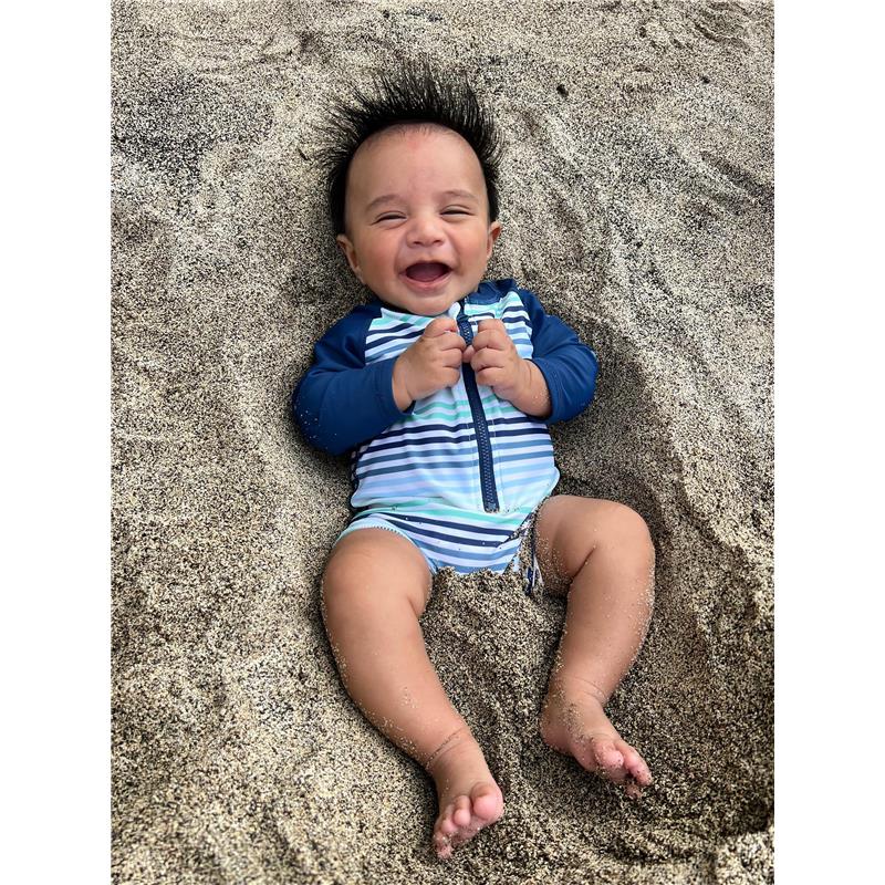 Baby boy lying in sand, wearing a striped long sleeve one-piece swimsuit with a zipper, smiling joyfully.