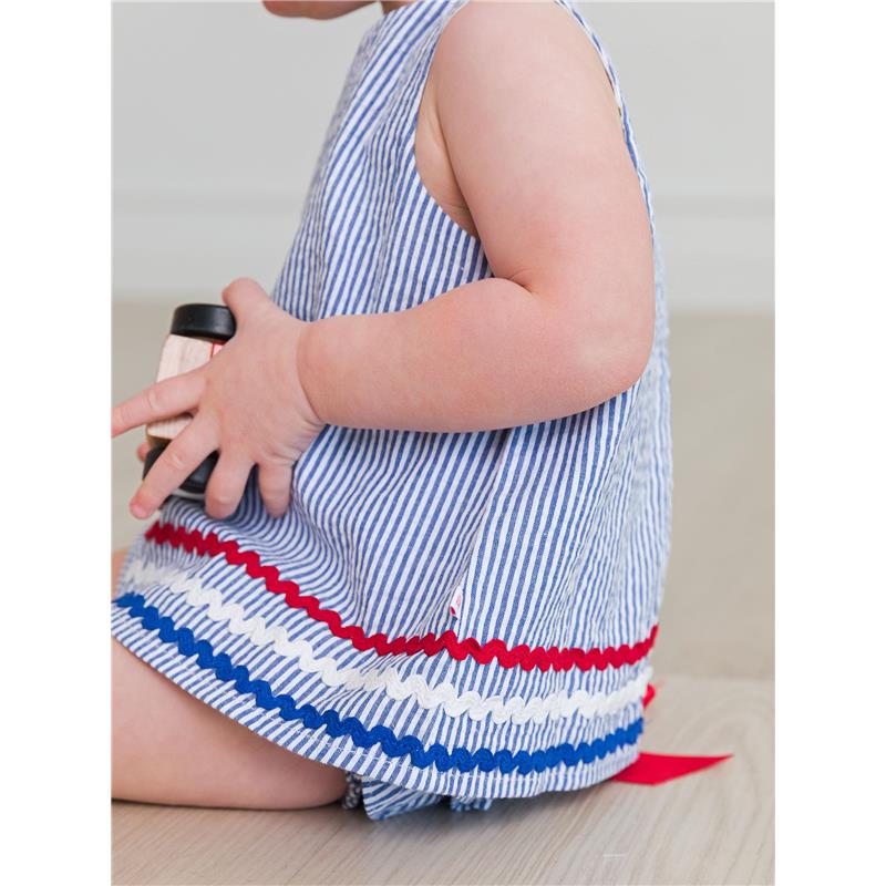 Baby girl wearing a blue seersucker swing top with red and blue ric rac trim, holding a toy.