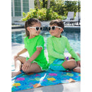 Two children relax by a pool, wearing bright neon honeydew long sleeve rash guards and sunglasses, sitting on a colorful towel.