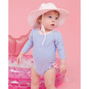 Baby wearing a periwinkle blue seersucker one-piece rash guard, pink and white striped sun hat, standing by a pink inflatable pool.