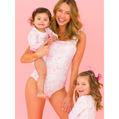 A woman holds a baby in a floral bubblegum pink ruffle one-piece swimsuit, smiling alongside a girl in a matching outfit.