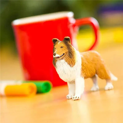 Lifelike Collie figurine by Safari Ltd. stands on a table beside a red mug and colored markers.