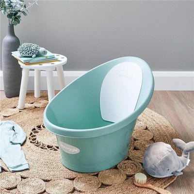 Newborn baby bath by Shnuggle in eucalyptus color, featuring a white support, with nearby toys and a blue garment on a woven rug.