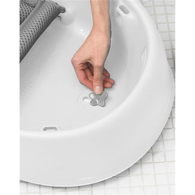 Hand adjusting the gray flower-shaped stopper in the bottom of the Skip Hop Moby Baby Bath Tub, which is white and designed for infants.
