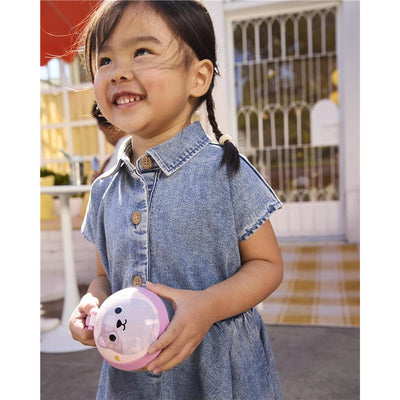 Girl with braided hair smiles while holding a pink cat-themed Skip Hop Baby Snack Container in a casual outdoor setting.