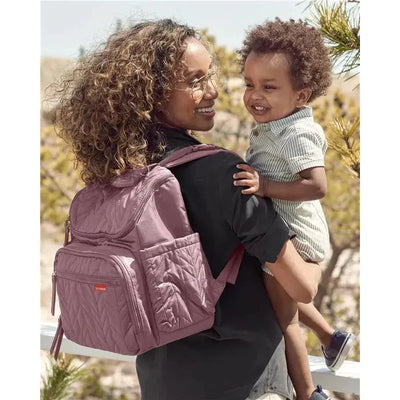 A woman smiles while holding a child on her hip, wearing a mauve diaper backpack with quilted detailing.