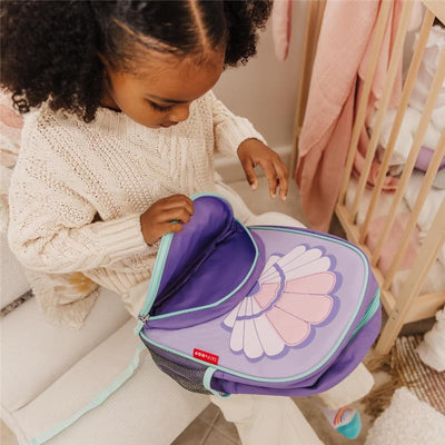 Little girl in a cozy sweater opening a Skip Hop Seashell backpack with a colorful design, sitting on a chair.