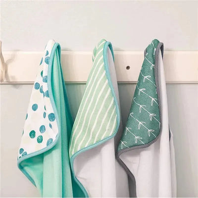 Three soft terry hooded towels in various designs: green dots, striped mint, and green with white leaf patterns, hanging on a hook.