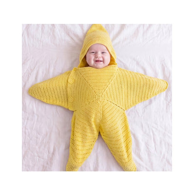 Smiling baby wearing a yellow hand-crocheted starfish jumpsuit with a matching hood, lying on a white blanket.