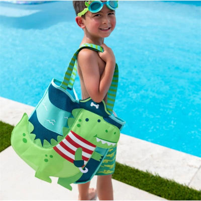A smiling boy in swim trunks holds a green dinosaur-themed beach tote near a pool, ready for fun.