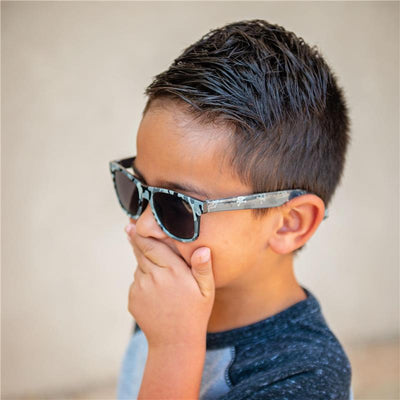 Boy with short hair wearing Stephen Joseph Flower sunglasses, covering his mouth with a hand, against a neutral background.