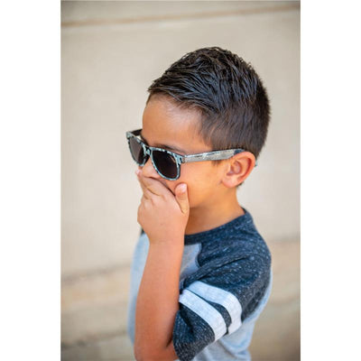 A boy wearing Stephen Joseph sunglasses with a patterned frame, covering his mouth in a playful pose.