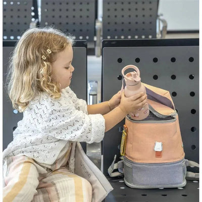 A young girl in a white sweater is placing a pink water bottle into a coral pink Stokke Jetkids backpack while seated.